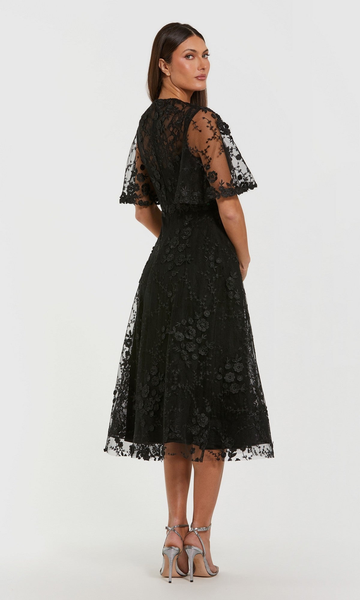 Woman in a Mac Duggal 69036 midi wedding guest dress with floral embroidery and short sheer sleeves, standing in heels and facing slightly away from the camera.